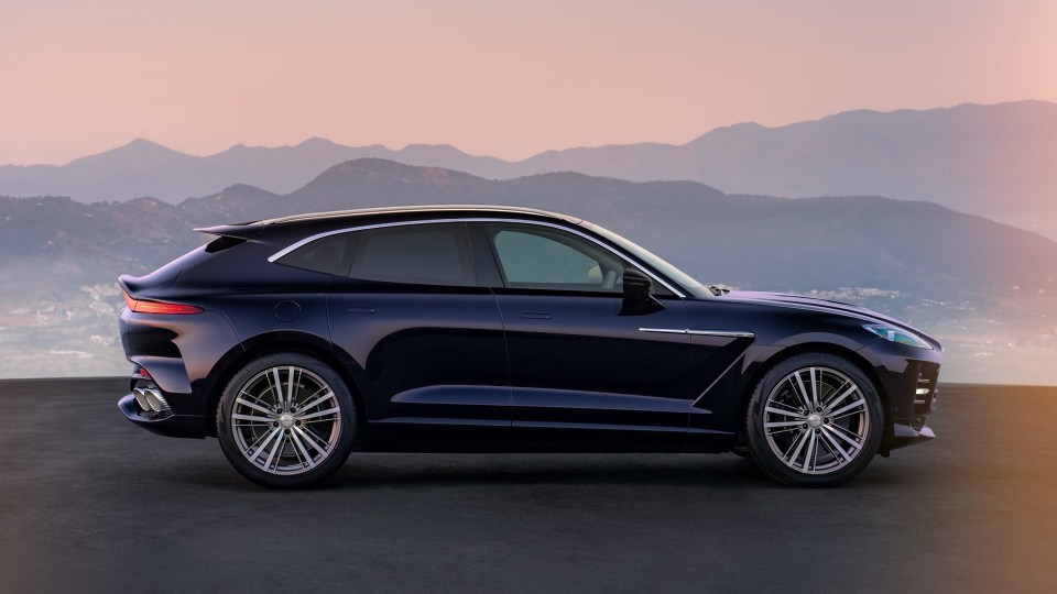 Dark blue luxury SUV, Aston Martin DBX, parked with a mountain range and sunset sky in the background.