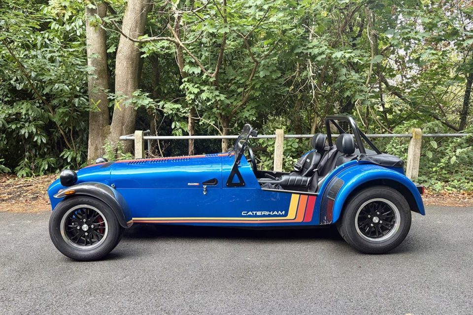 Blue Caterham Seven sports car with retro stripes parked on asphalt, surrounded by green trees.