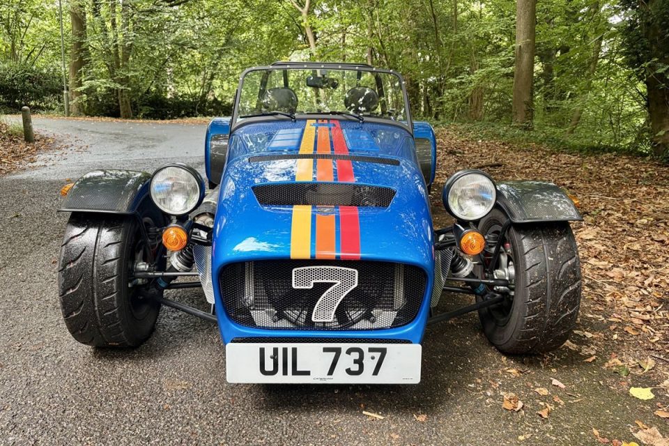 Front view of a blue Caterham Seven sports car with orange and red racing stripes, parked on an asphalt road.