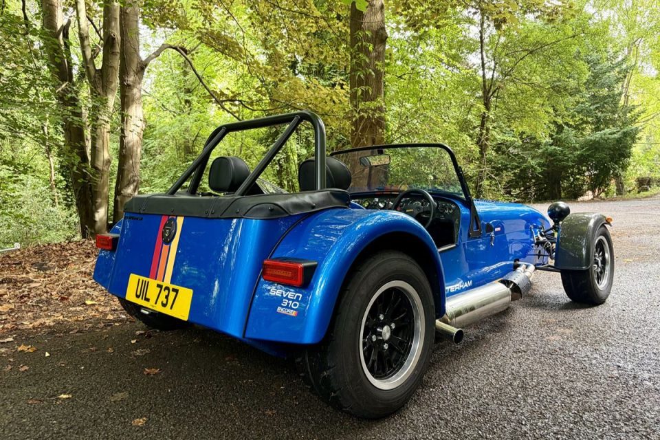 Blue Caterham Seven 310 Encore sports car with racing stripes, parked on an asphalt road in a wooded area.