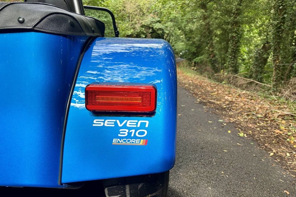 Rear view of a blue Caterham Seven 310 Encore sports car on a winding road lined with trees.