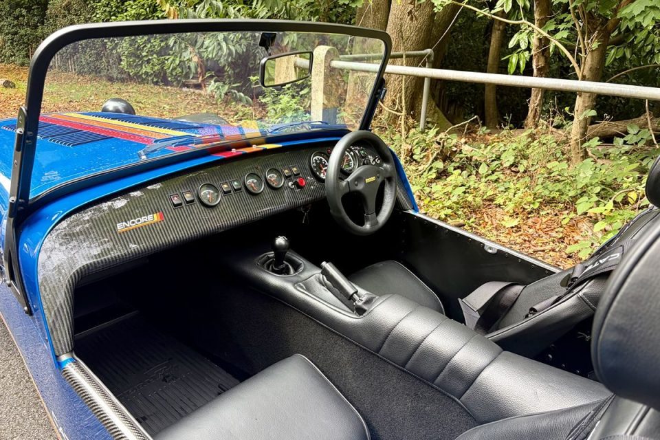 Interior of a blue Caterham sports car with carbon fiber dashboard, black leather seats, and Encore branding.
