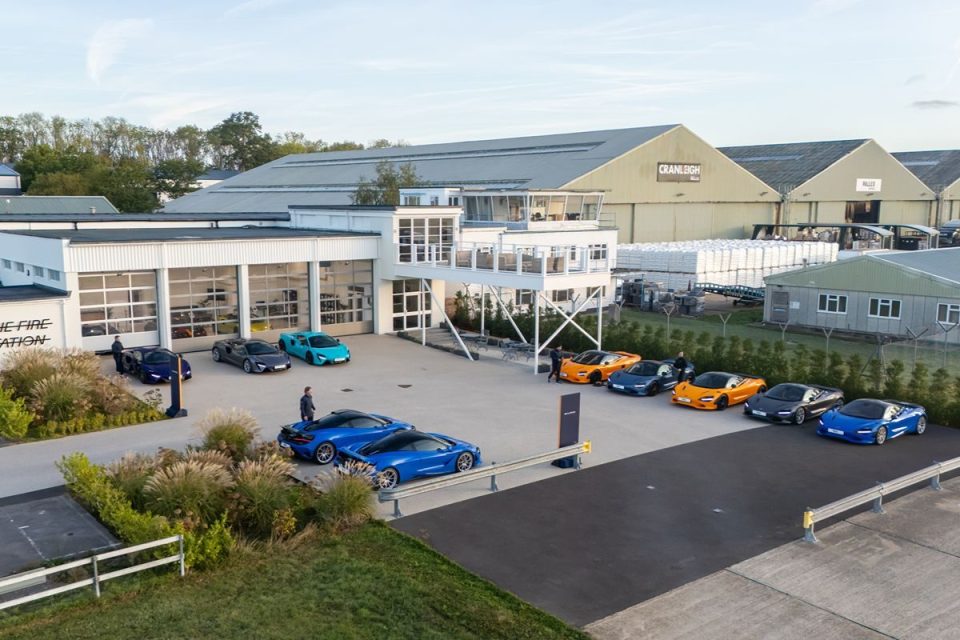 Modern industrial complex with several McLaren sports cars parked in front, including models in blue, orange, and grey.