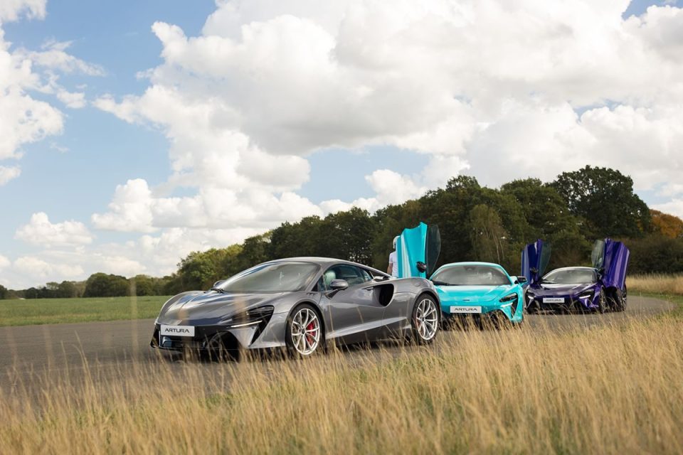 Three McLaren Artura supercars in grey, blue, and purple with open dihedral doors on a track with grass and trees.
