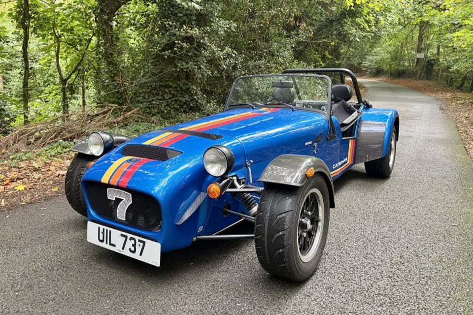Blue Caterham Seven sports car with racing stripes on a paved road in a wooded area