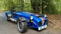 Blue Caterham sports car with racing stripes, number 7, parked on an asphalt road in a wooded area