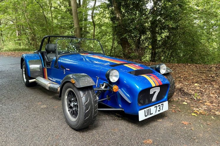 Blue Caterham sports car with racing stripes, number 7, parked on an asphalt road in a wooded area