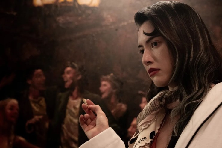 Woman with dark hair and red lipstick holding a small object, with blurred figures in the background.
