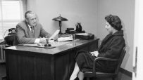 Man in suit conducting a job interview with a woman in a patterned coat, 1950s office setting.