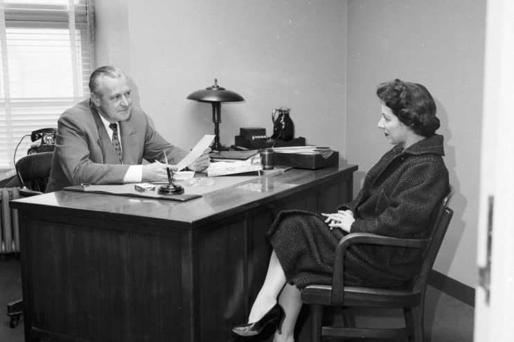 Man in suit conducting a job interview with a woman in a patterned coat, 1950s office setting.