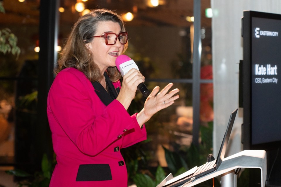 Kate Hart, CEO of Eastern City, speaking at a podium with a microphone, wearing a bright pink jacket