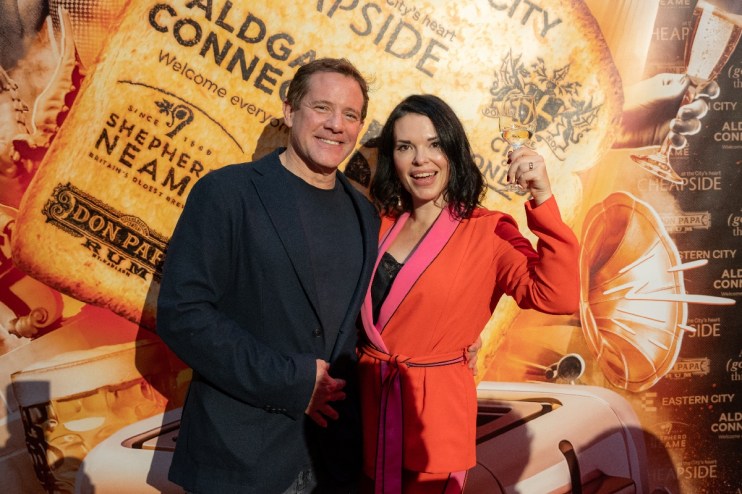 Man and woman smiling, woman holding wine glass, against a branded backdrop for Libby Toast wine column.