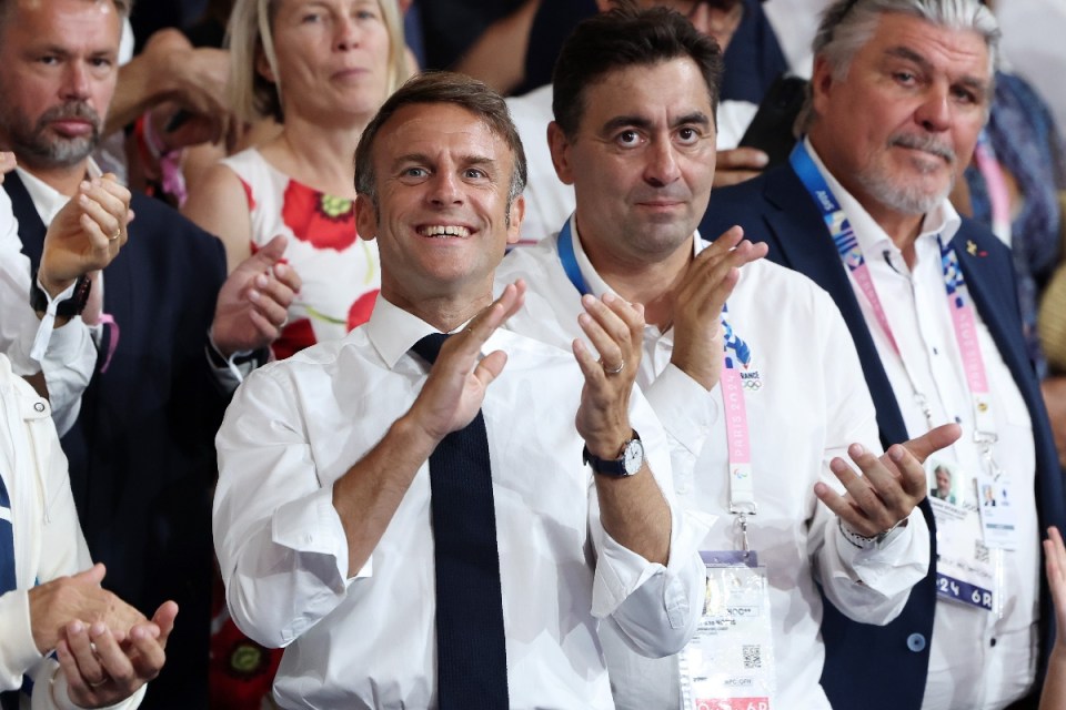 Emmanuel Macron smiling and clapping, surrounded by officials, at a Paris 2024 Olympic event.