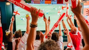 Football fans in a pub cheering with England flags, inflatable sticks, and drinks, watching a World Cup game.