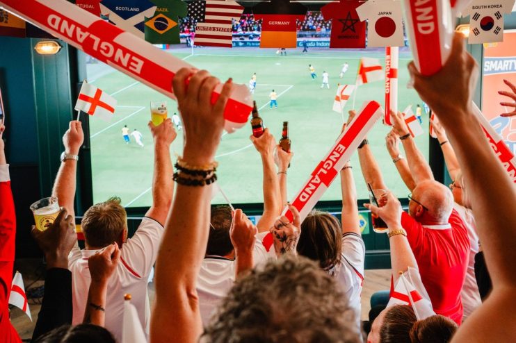 Football fans in a pub cheering with England flags, inflatable sticks, and drinks, watching a World Cup game.