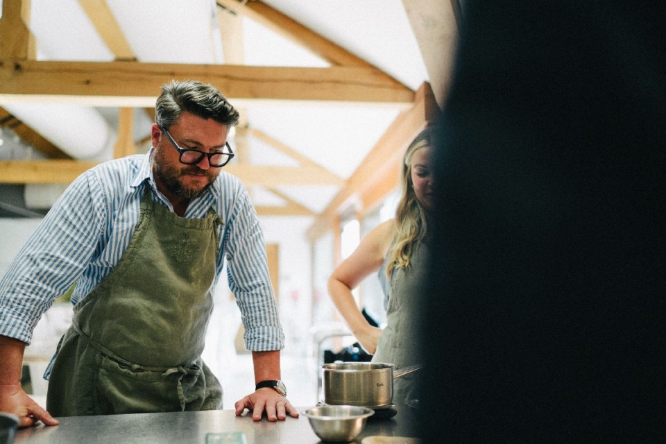 Adam looking down, wearing glasses and a chefs apron, with a woman in an apron and cooking pot nearby.