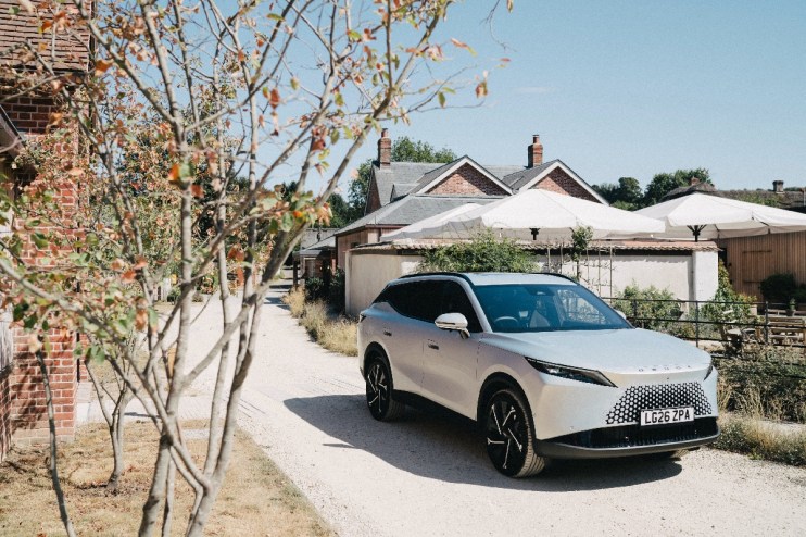 Silver Omoda 7 SUV parked on a gravel path next to brick buildings and white patio umbrellas