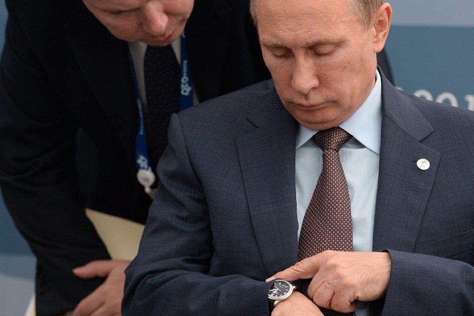 Vladimir Putin in a dark suit, light blue shirt, and patterned tie, checking his wrist watch.