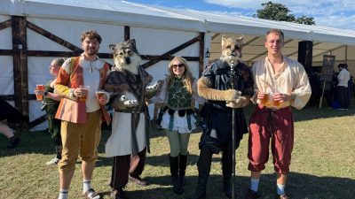 People in Renaissance Fair costumes with furry animal masks holding drinks outdoors.