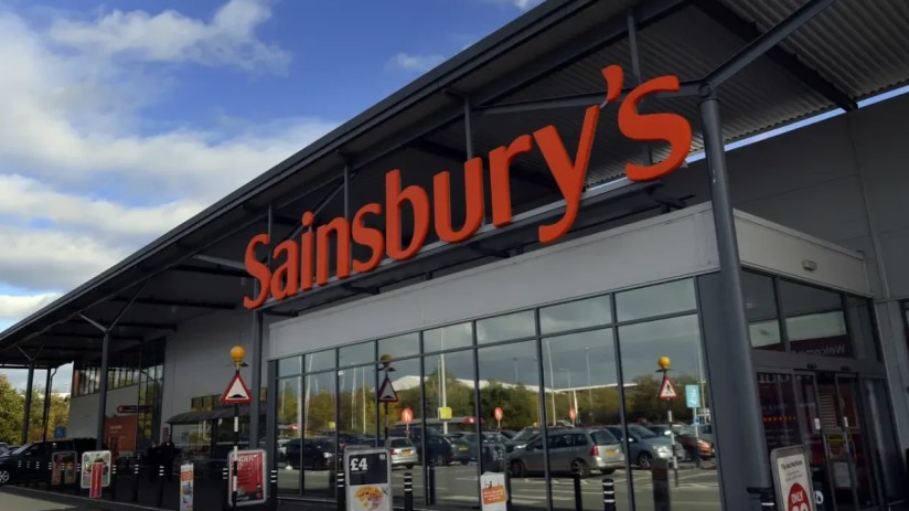 Sainsburys supermarket exterior with prominent orange logo, large windows reflecting parked cars, and a blue sky with clouds