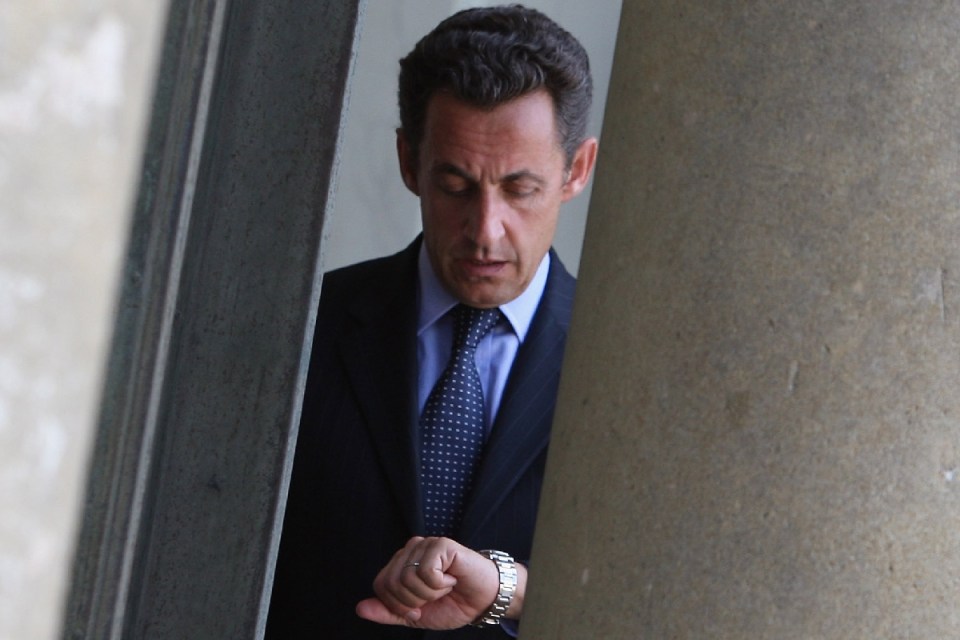 Nicolas Sarkozy checking his watch while peeking from behind a column, wearing a suit and spotted tie