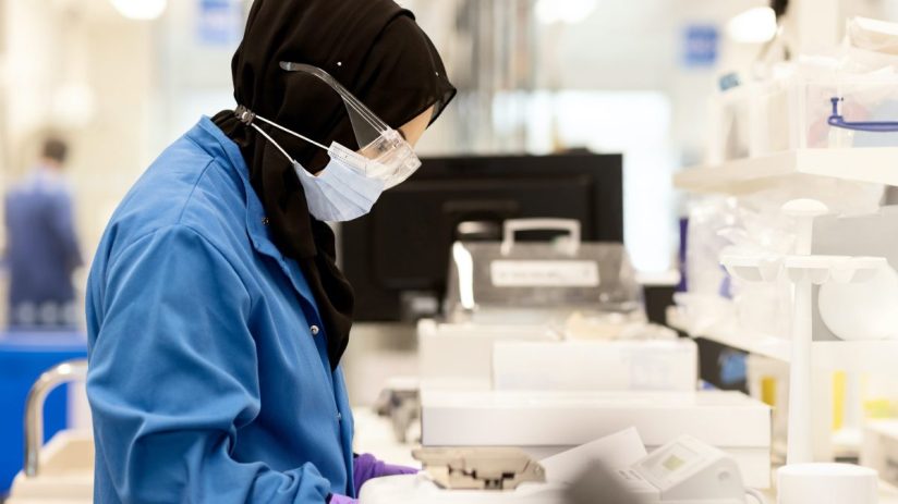 Scientist in hijab, lab coat, mask, and safety glasses inspecting paperwork in a clean laboratory.
