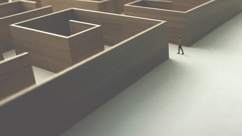 Small figure in a tan coat walks on a white path next to a large, wooden maze, symbolizing business challenges.