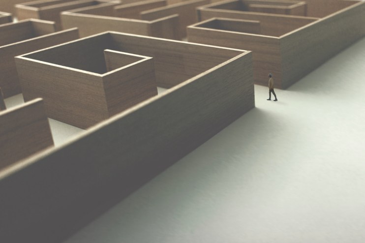 Small figure in a tan coat walks on a white path next to a large, wooden maze, symbolizing business challenges.