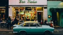 A teal vintage car parked in front of Bar Italia in Soho, with people on the sidewalk and inside the cafe.