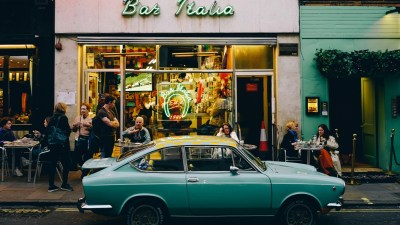A teal vintage car parked in front of Bar Italia in Soho, with people on the sidewalk and inside the cafe.