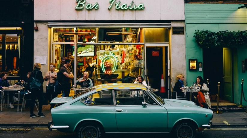 A teal vintage car parked in front of Bar Italia in Soho, with people on the sidewalk and inside the cafe.