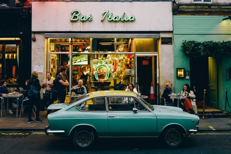 A teal vintage car parked in front of Bar Italia in Soho, with people on the sidewalk and inside the cafe.