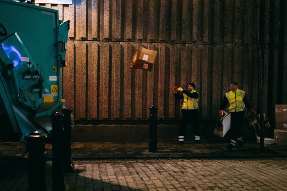 Soho bin men in hi-vis vests disposing of refuse into a large turquoise bin lorry at night