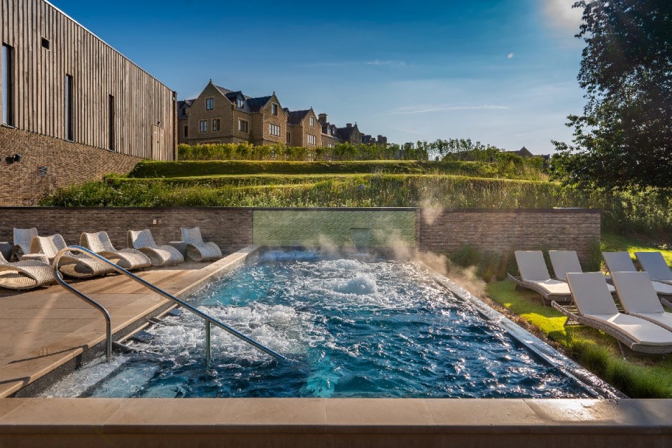 Outdoor hydrotherapy pool at South Lodge Spa with steam rising, surrounded by lounge chairs and hotel buildings in the bac...