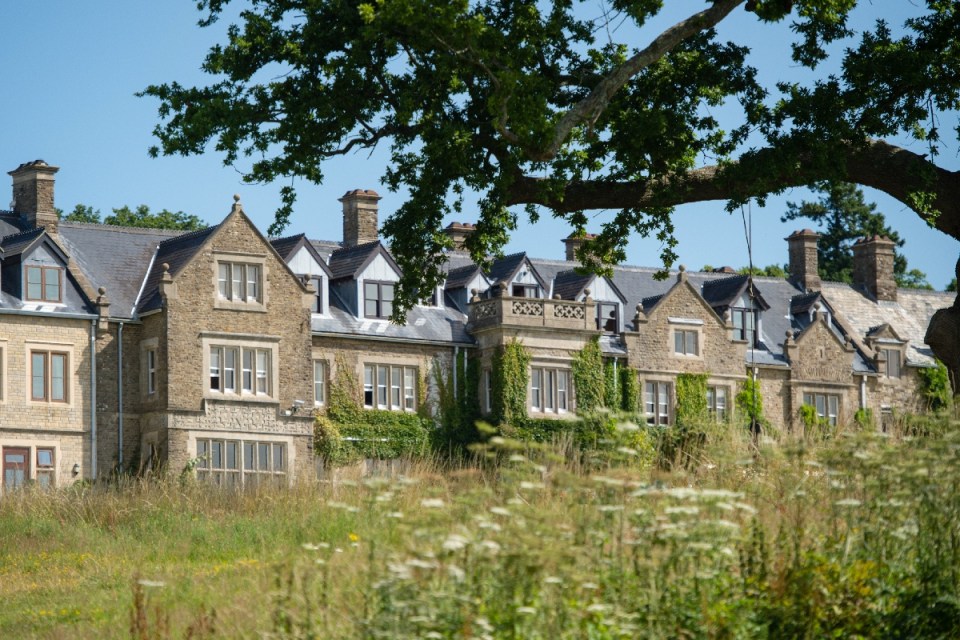 Historic South Lodge building with stone facade, slate roofs, and climbing ivy, seen through a field of wildflowers.