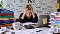 Stressed woman surrounded by tax forms, binders, and a TAX mind map on a whiteboard