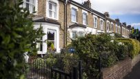 Row of traditional London terraced houses with bay windows, brick facades, and front gardens, under a blue sky.
