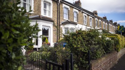 Row of traditional London terraced houses with bay windows, brick facades, and front gardens, under a blue sky.