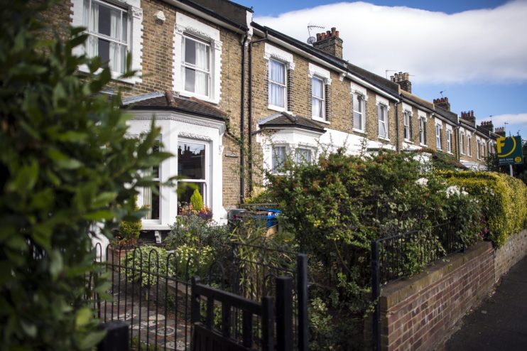 Row of traditional London terraced houses with bay windows, brick facades, and front gardens, under a blue sky.