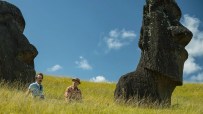 Two men in casual attire sit in tall green grass between large Moai statues under a bright blue sky.