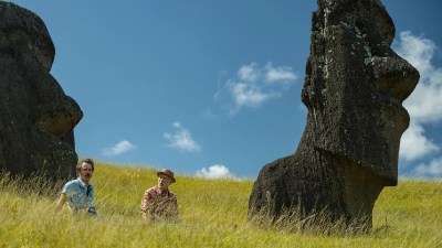 Two men in casual attire sit in tall green grass between large Moai statues under a bright blue sky.