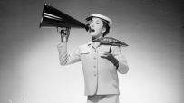 Woman in a skirt suit and hat speaking into a megaphone, gesturing with her free hand.