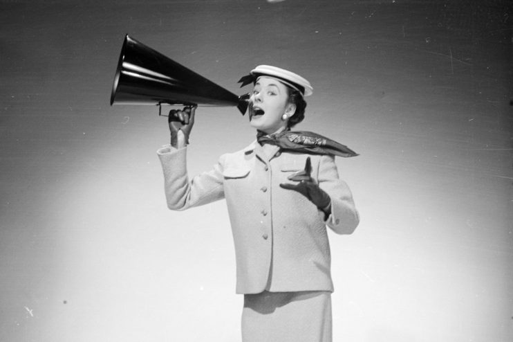 Woman in a skirt suit and hat speaking into a megaphone, gesturing with her free hand.